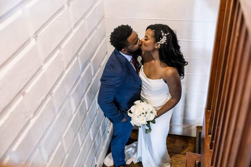A bride and groom kissing in a stairwell, her bouquet between them