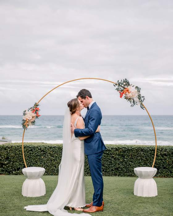 A bride and groom under a circular floral arch on a lawn above the ocean