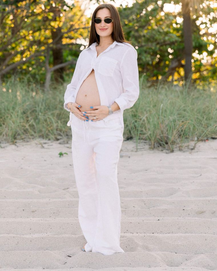 An expecting mother in white linen on the sand at golden hour