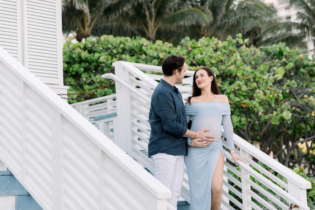 An expecting couple on a white beach staircase at golden hour