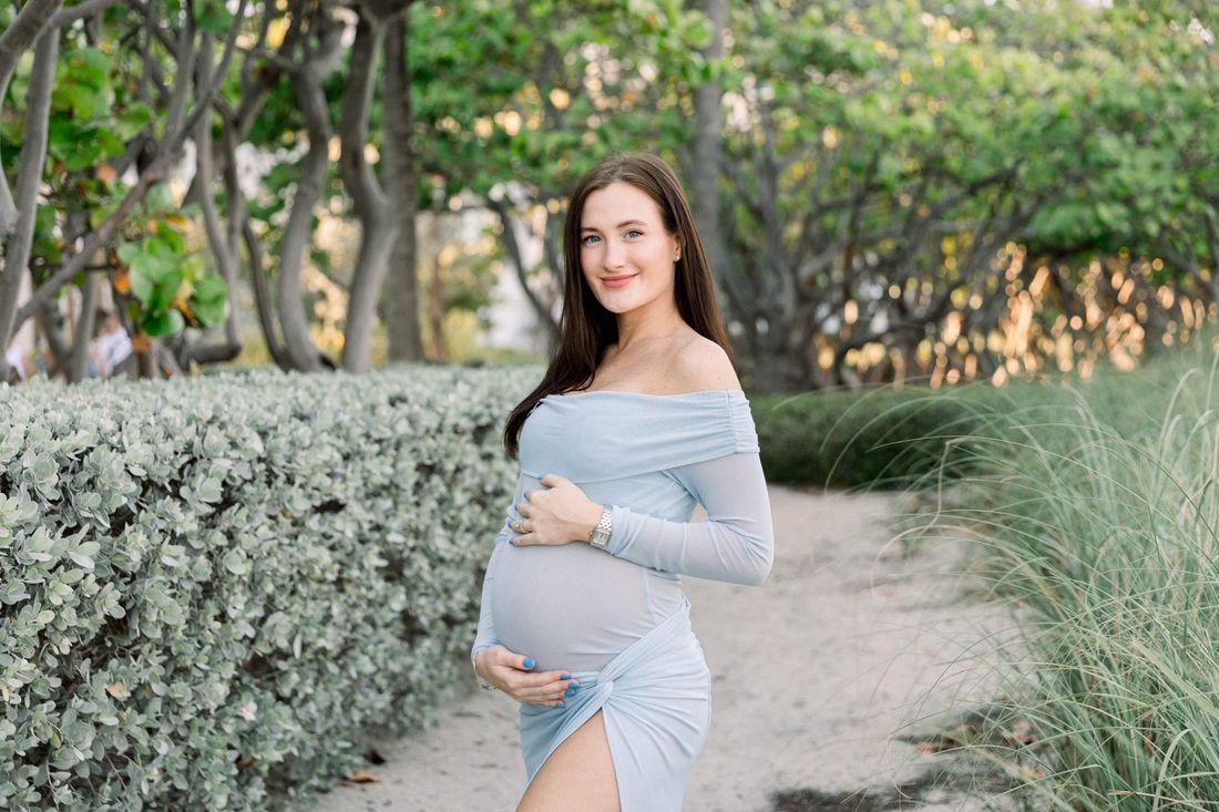 An expecting mother in a pale blue dress on a beach path at sunset